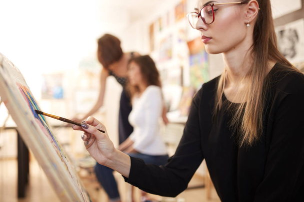 Nice young girl in glasses dressed in black blouse and jeans sits at the easel and paints a picture in the drawing school - Foto, Imagen