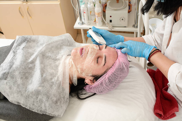 Jovem mulher no salão de beleza fazendo ultra-som descascamento e limpeza facial procedimento. - Foto, Imagem