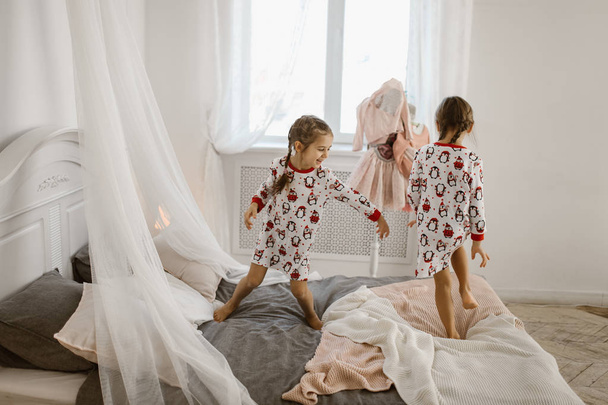 Dos niñas en pijama se divierten saltando en una cama en un acogedor dormitorio iluminado por el sol
 - Foto, imagen