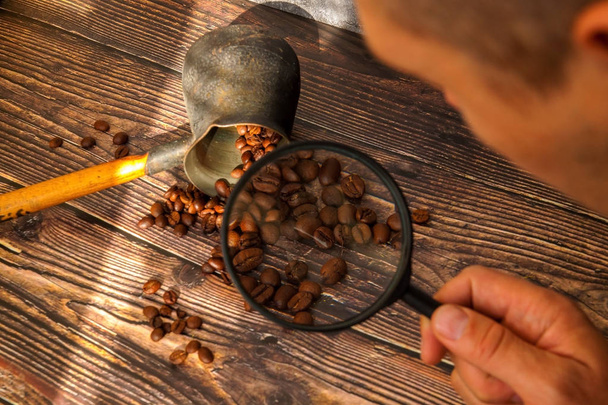 Man examines coffee beans through a magnifying glass. man's hand keeps magnifying glass over the coffee beans. - Fotó, kép