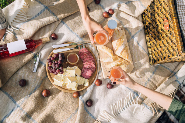 A healthy picnic on summer holidays with freshly baked bread, cheese and salami is laid out on a brown plaid. Girls drink multifruit juice in the park. - Foto, Imagem
