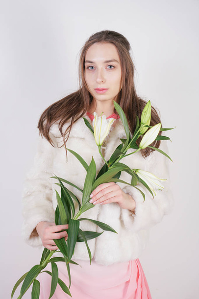 ženská dívka v bílém kožešním kabátě s větví lilií na pozadí studia - Fotografie, Obrázek