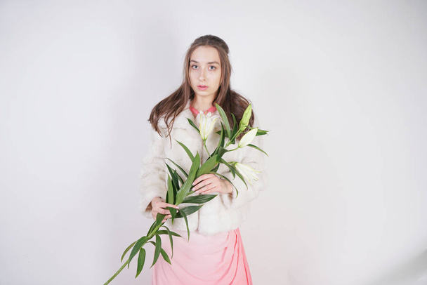 feminine girl in a white fur coat with a branch of lilies in the background in the Studio - Fotografie, Obrázek