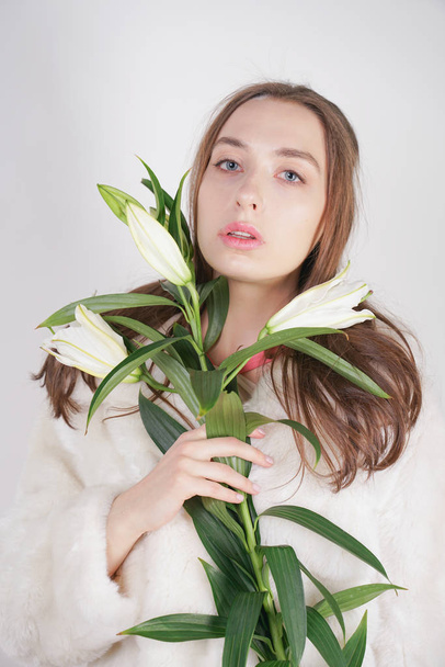 feminine girl in a white fur coat with a branch of lilies in the background in the Studio - Foto, Imagen
