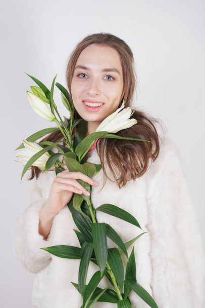 feminine girl in a white fur coat with a branch of lilies in the background in the Studio - Photo, Image