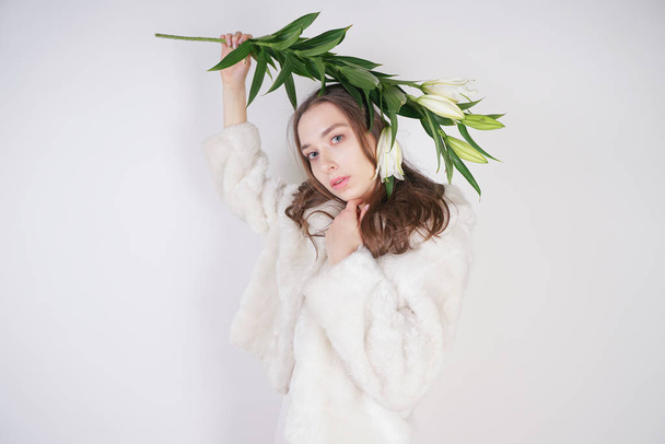 feminine girl in a white fur coat with a branch of lilies in the background in the Studio - Fotoğraf, Görsel