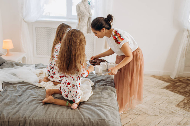 Young beautiful mother is bringing cocoa with Marshmallows and cookies to her daughters sitting on the bed in the full of light cozy room - Φωτογραφία, εικόνα