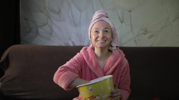 Woman watching a late night movie at TV, eating popcorn. Bathrobe, facial mask - Footage, Video