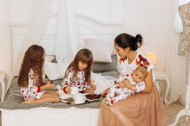 Young mother with her tiny baby sits o the bed with her two  daughters dressed  in pajamas eating cookies with  cocoa with marshmallows - Foto, imagen
