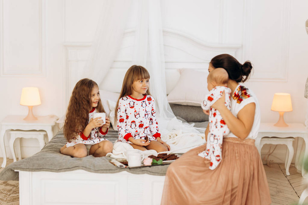 Young mother with her tiny baby sits o the bed with her two  daughters dressed  in pajamas eating cookies with  cocoa with marshmallows - Foto, imagen