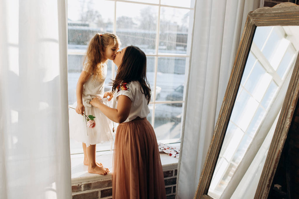 Young mother kisses her little daughter standing on the windowsill next to the mirror in the full of light cozy room - Valokuva, kuva