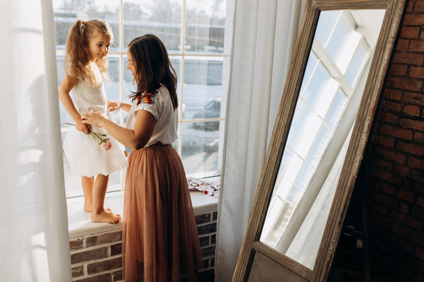 Young mother holds her little daughter standing on the windowsill next to the mirror in the full of light cozy room - Fotografie, Obrázek