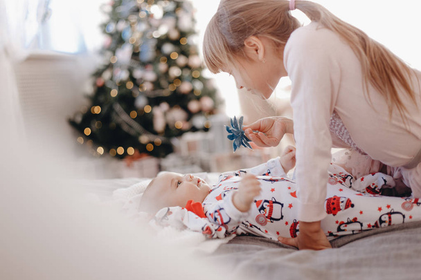 Little girl dressed in pajama is looking at her tiny brother lying on the bed in the cozy room with New Years tree - Φωτογραφία, εικόνα
