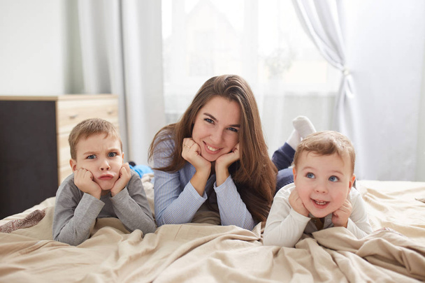 Joyeux jeune mère vêtue de pyjama bleu clair couche avec ses deux petits fils tenant leurs mains sous leurs mentons sur le lit avec couverture beige dans la chambre avec grande fenêtre
 - Photo, image