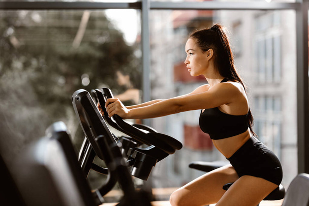 Young athletic girl with brown hair dressed in black sports top and shorts works out on the exercise bike in the gym - Foto, imagen