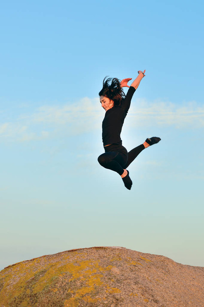 young woman doing gymnastic jump on a rock against a blue sky - Fotó, kép