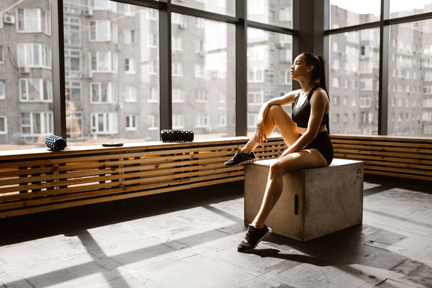 Charming slander girl dressed in black sports top and shorts is sitting on a wooden box in the sunlight in front the window in the gym - Foto, Bild