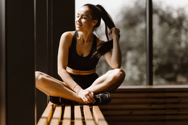 Slender dark-haired girl dressed in black sports top and shorts is sitting in lotus pose on a wooden window sill in the gym - Photo, image