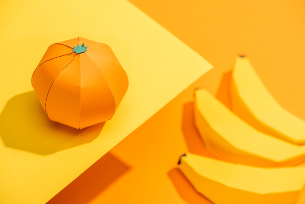 Selective Focus of origami tangerine on yellow paper with cardboard bananas on orange - Photo, Image
