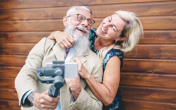幸せな高齢者カップルは、ジンバル携帯電話屋外でビデオを録画 - 新しいトレンドのスマートフォンアプリを楽しむ成熟したファッションの人々 - 木製の背景 - 高齢者のライフスタイルと技術コンセプト - 写真・画像