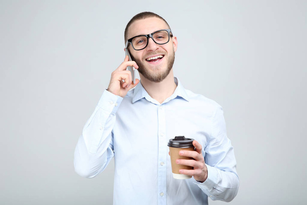 Young man in glasses with smartphone and paper cup on grey backg - Foto, immagini