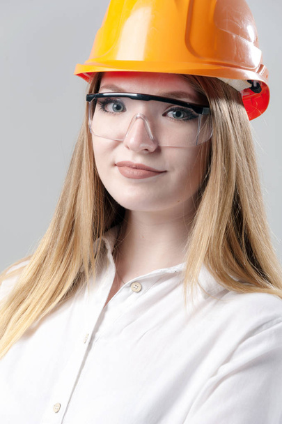 Portrait of a young attractive woman with blond hair in orange helmet on a neutral gray background. - Foto, Imagen