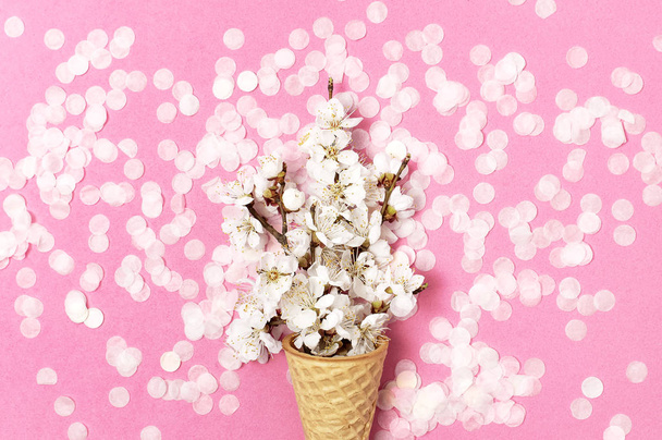 Waffle cone with bouquet of spring white flowers and round confetti on pastel pink background. Festive holiday background, concept of party, birthday. Creative spring minimal style Flat lay top view - Valokuva, kuva
