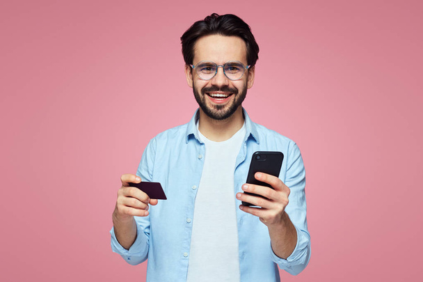 Привлекательный красивый мужчина улыбается, держа в руках пластиковую кредитку и мобильный телефон, изолированные на розовом фоне
. - Фото, изображение