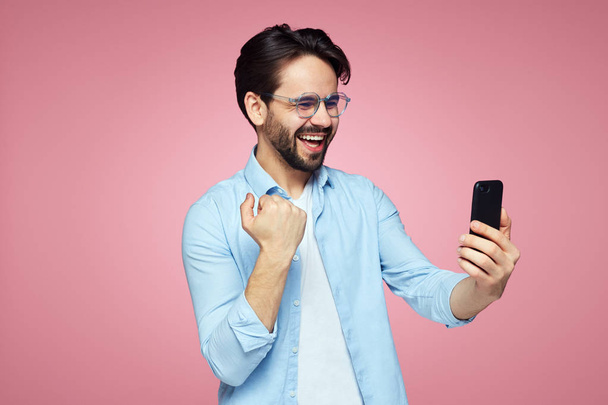 Jeune excité mâle en chemise bleue tenant téléphone portable et montrant geste gagnant avec poing debout sur fond de studio rose
 - Photo, image