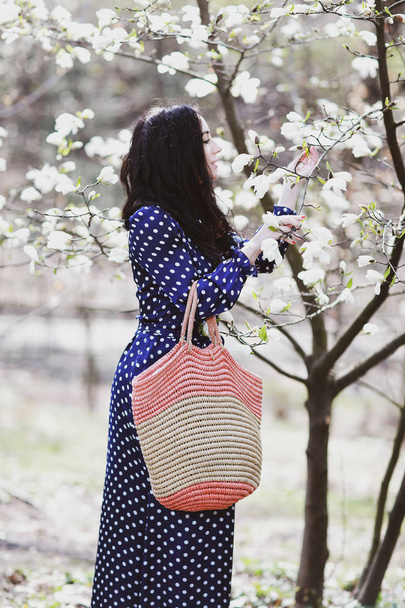 beautiful brunette woman in a blue dress and a knitted bag near magnolia blooming - Zdjęcie, obraz