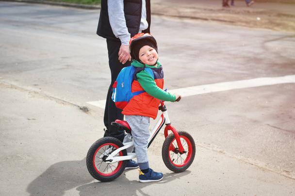 Een kleine coole jongen in een helm en een rood vest staat met zijn vader in de buurt van de weg op een Balance Bike met een blauwe rugzak op zijn rug. - Foto, afbeelding