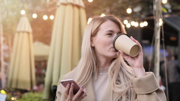 Gorgeous young woman with cup of coffee and mobile phone in city street. Stylish happy hipster girl using modern smartphone device. - Fotografie, Obrázek