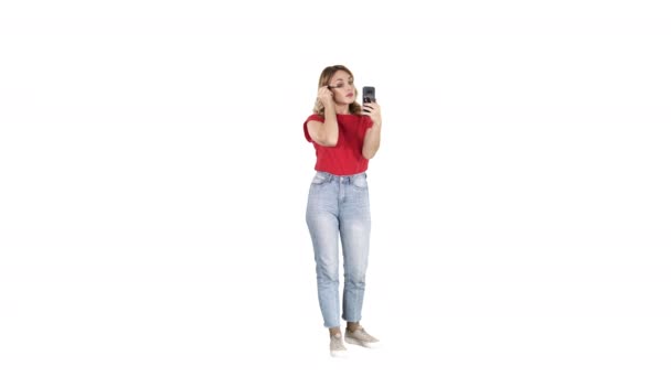 Gyönyörű fiatal nő használ szempillaspirál és keres az ő smartphone fehér háttér. - Felvétel, videó