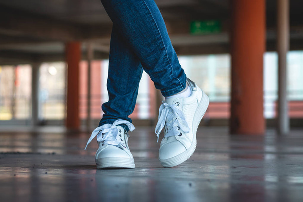 Woman with white sneakers standing in public parking garage. Fashion concept. Slim female legs wearing jeans and sport shoes in parking lot.  - 写真・画像