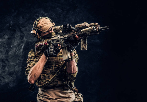 Элитный отряд, солдат спецназа в камуфляжной форме держит штурмовую винтовку с лазерным прицелом и целится в цель. Студийное фото у темной стены
   - Фото, изображение