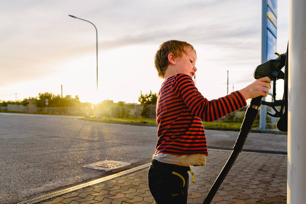 Kind hält den Schlauch einer Zapfsäule bei Sonnenuntergang an einer Tankstelle - Foto, Bild