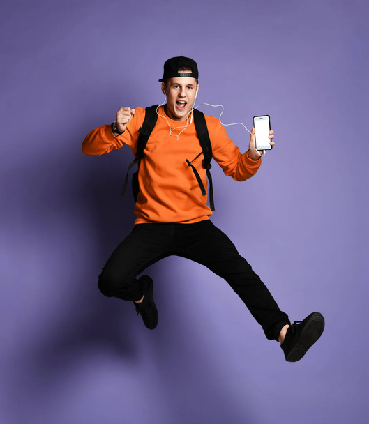young student man in casual wear with backpack and headphones is listening to music using a smartphone and smiling while jumping - Foto, immagini