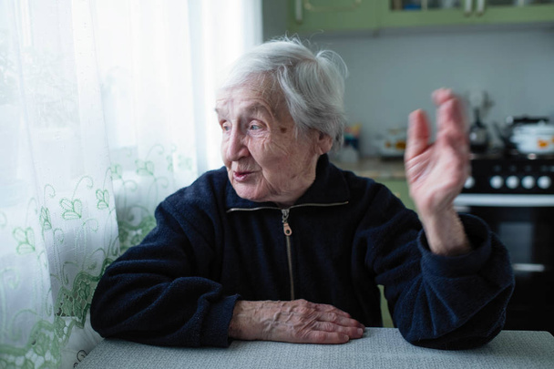 De oude vrouw spreekt expressief zittend aan tafel. - Foto, afbeelding