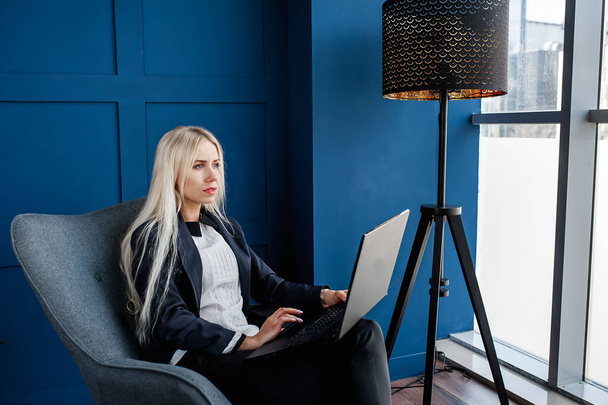 Elegancka biznesmen siedzący w fotelu i pracujący na laptopie w niebieskim wnętrzu  - Zdjęcie, obraz