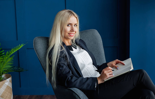 junge blonde Frau hält Notizbuch und sitzt im grauen Sessel  - Foto, Bild