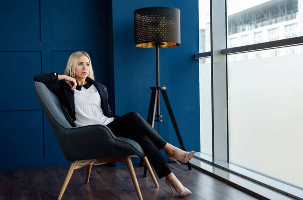 Elegant businesswoman sitting in armchair in blue studio  - Фото, изображение