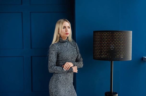 Young blonde woman in grey stylish dress posing in blue studio interior - Zdjęcie, obraz