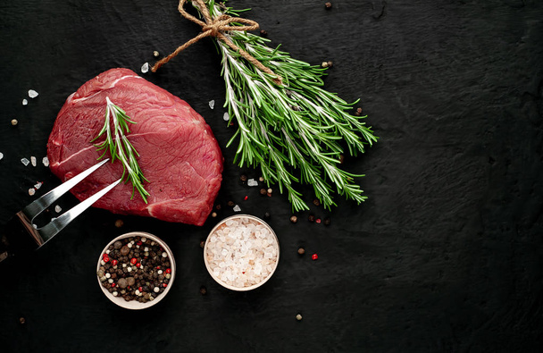 Raw steak with green rosemary twigs and spices on black stone background  - Photo, Image