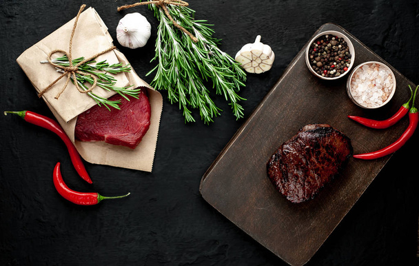 Grilled steak and raw meat wrapped in craft paper on black stone background - Фото, зображення