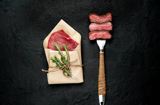 Filete en rodajas apuñalado en tenedor y carne cruda envuelta en papel sobre fondo de piedra negra
  - Foto, imagen