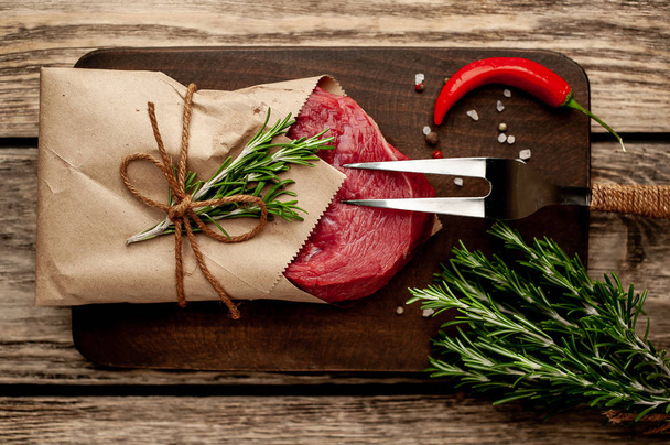 Steak de boeuf cru enveloppé dans du papier artisanal avec brindilles de romarin sur planche à découper en bois
 - Photo, image