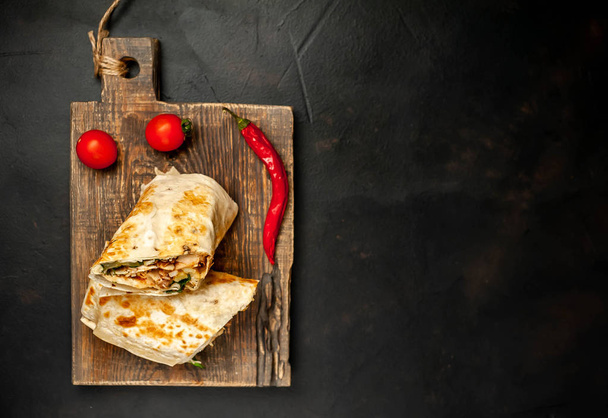 Traditional mexican burrito wrapped with chicken and vegetables served with ripe tomatoes on wooden chopping board  - Photo, Image