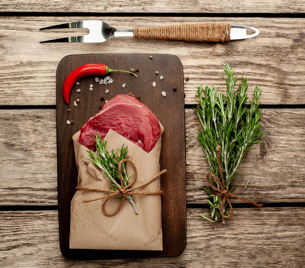 Steak de boeuf cru enveloppé dans du papier artisanal avec brindilles de romarin sur planche à découper en bois
 - Photo, image
