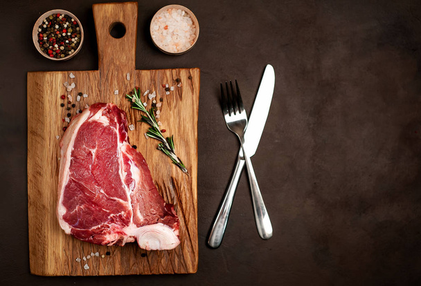 Raw fat steak with spices and rosemary twig on wooden chopping board over stone background - Photo, Image