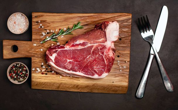 Tlustý steak s kořením a rozmarní větvičku na dřevěné sekací desce nad kamenným pozadím - Fotografie, Obrázek